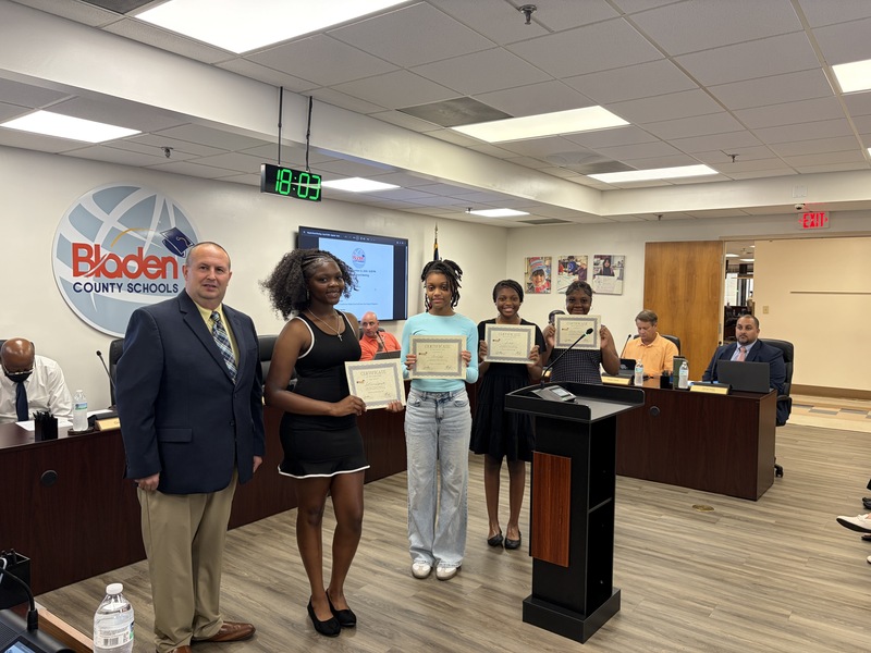 Four middle school students stand holding certificates with the superintendent.