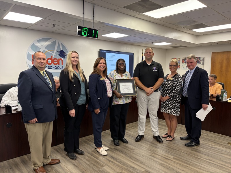 Adults stand together smiling with one employee holding a certificates of recognition in the middle.