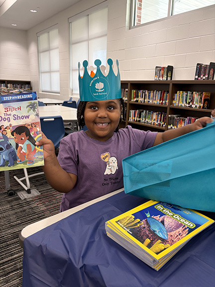 An elementary student stands smiling in the library holding a book.