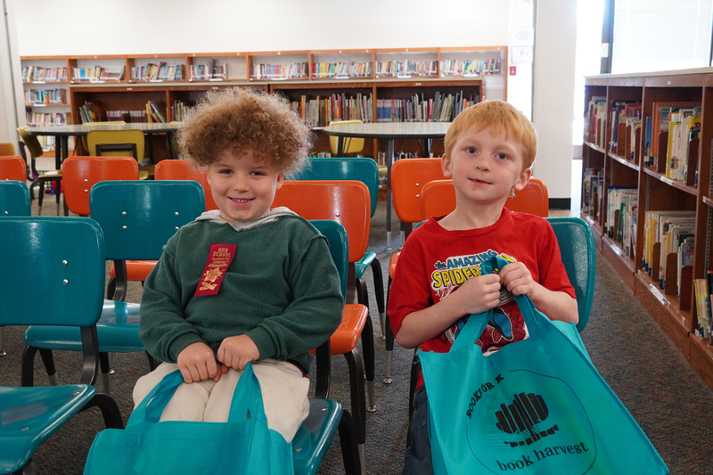 Two kindergarten boys sit in charis in a library holding teal bags.