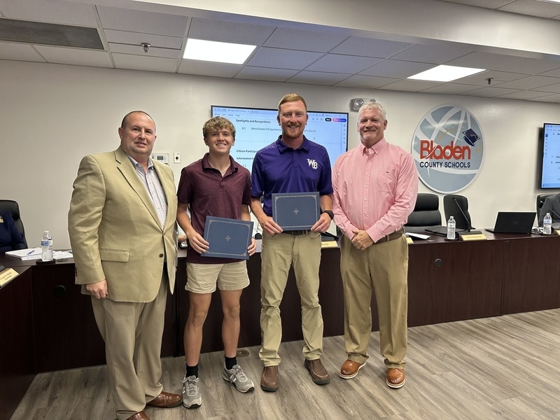 A student athlete and his coach stand at the front of a boardroom holding certificates, joined by two adult men in professional attire. The Bladen County Schools logo is displayed on the wall behind them.