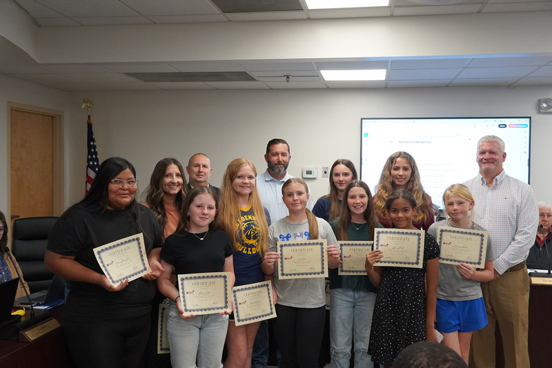 A group of middle school girls stand together with their coach, the superintendent and board chair. white background with flag in the corner