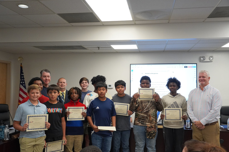 a gruop of middle school boys stand together holding certificates along with heir coarch, superintendent, and board chair. there is a white background and american flag in the corner.