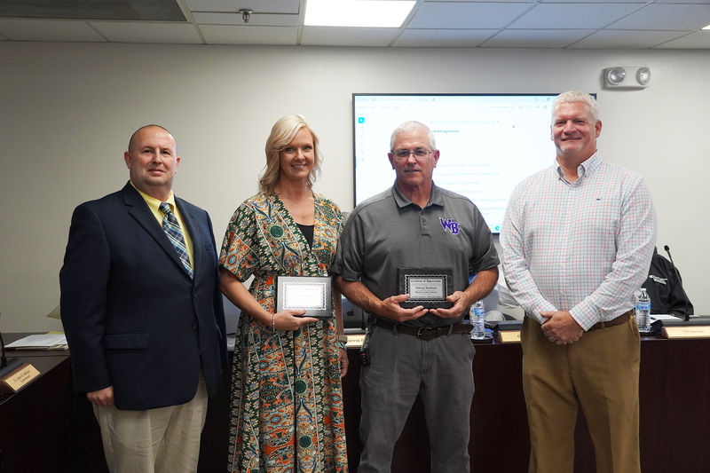 Two staff members stand between school leaders holding plaques of recognition during a Bladen County Schools board meeting. The woman wears a patterned dress, and the man beside her wears a gray West Bladen polo. Behind them, a presentation slide is displayed on a large screen.