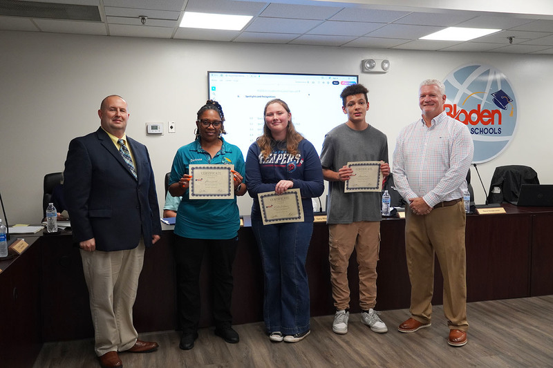 1 staff member and two students stand holding certificates between the superintendent and board chair. 