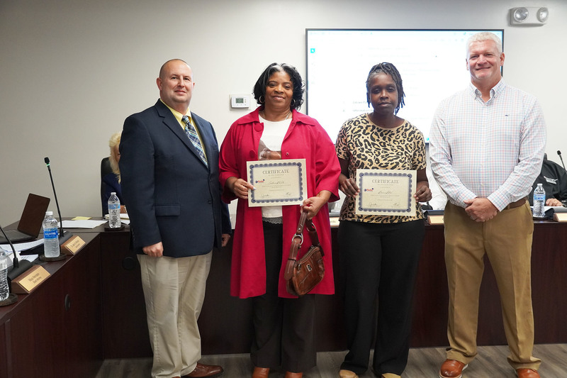 two staff members stand holding certificates between the superintendent and board chair