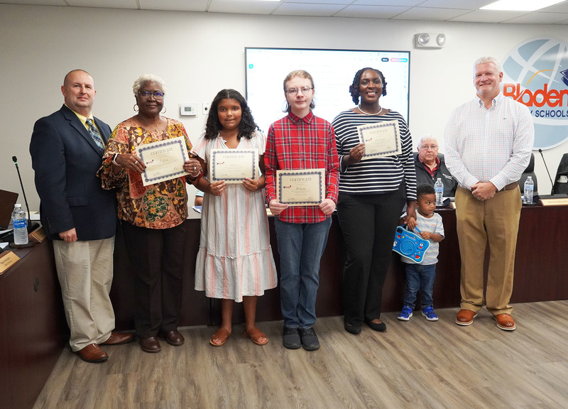 two students and two staff members stand holding certificates between the superintendent and board chair. one staff member also has a young child with her.