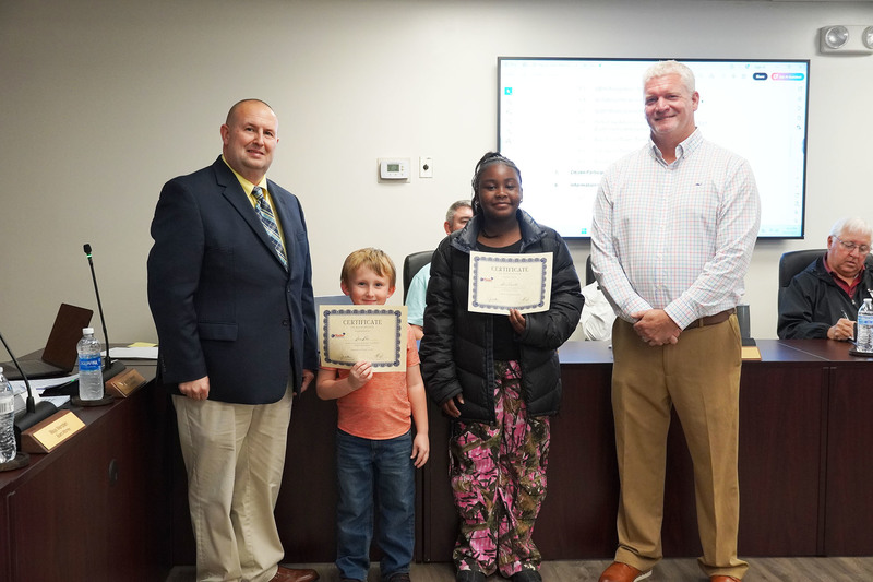 two students stand holding certificates between the superintendnet and board chair