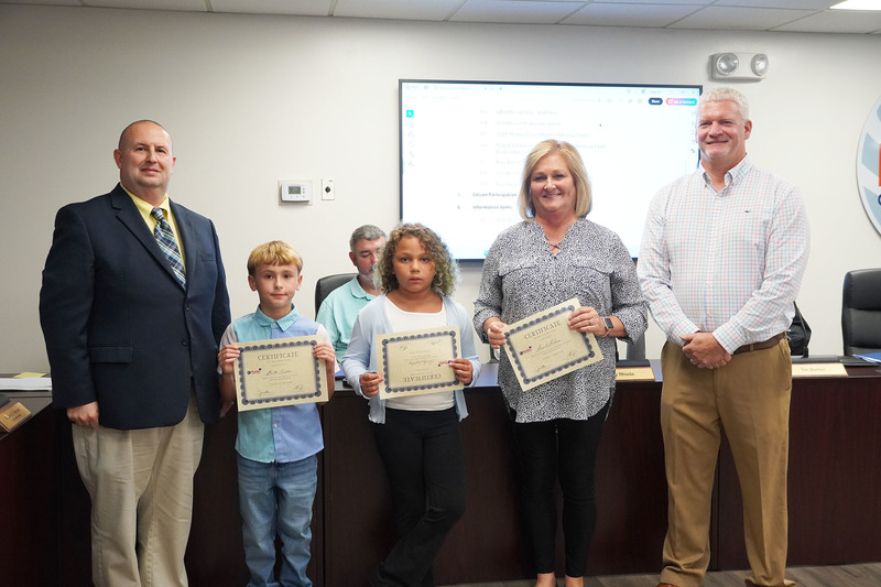 2 students and 1 staff member stand holding certificates between the superintendent and board chair.