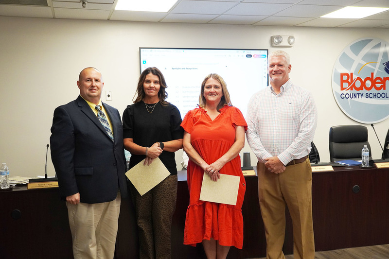 2 staff members standing holidng certificicates between the superintendent and board chair