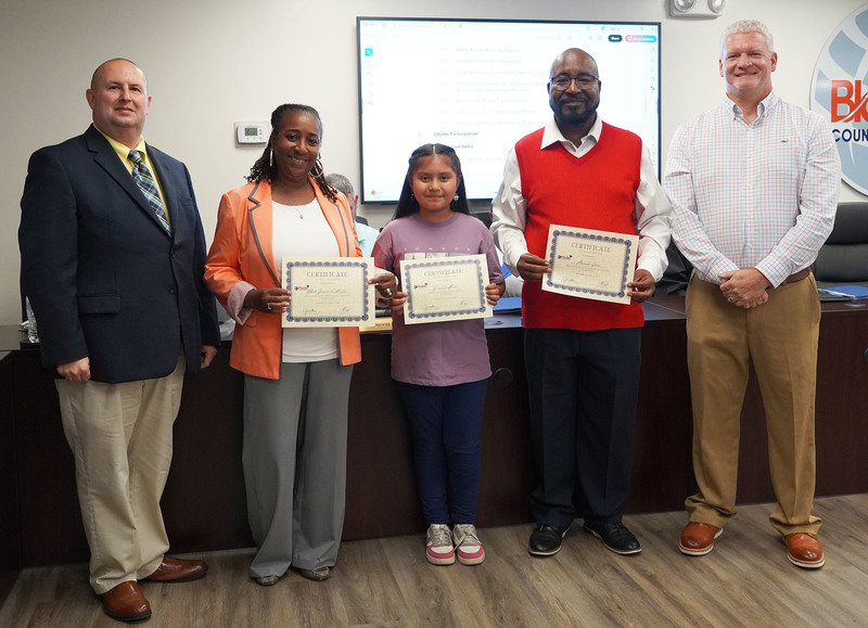 two staff members and a student stand together holding certificates between the superintendent and board chair