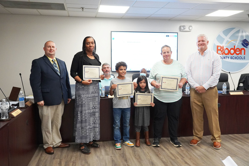two employees and two staffs stand between the superintendent and board chair while holding certificates of recognition.