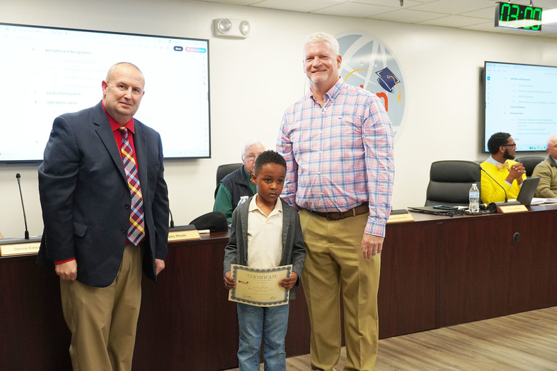 Young student holding a certificate stands with two administrators during a board meeting recognition.