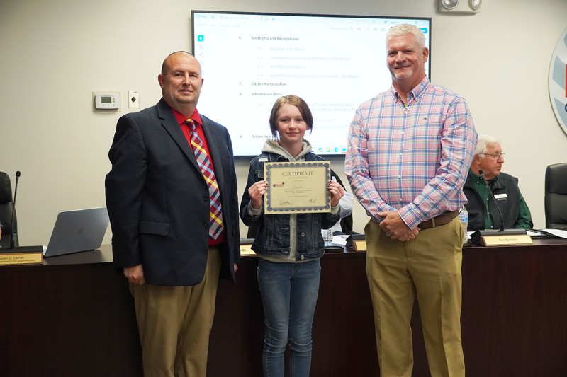 Student holding a certificate stands between two administrators during a recognition at a board meeting.