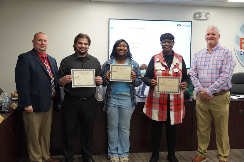 Two students and one staff member hold certificates during a recognition with district leaders.