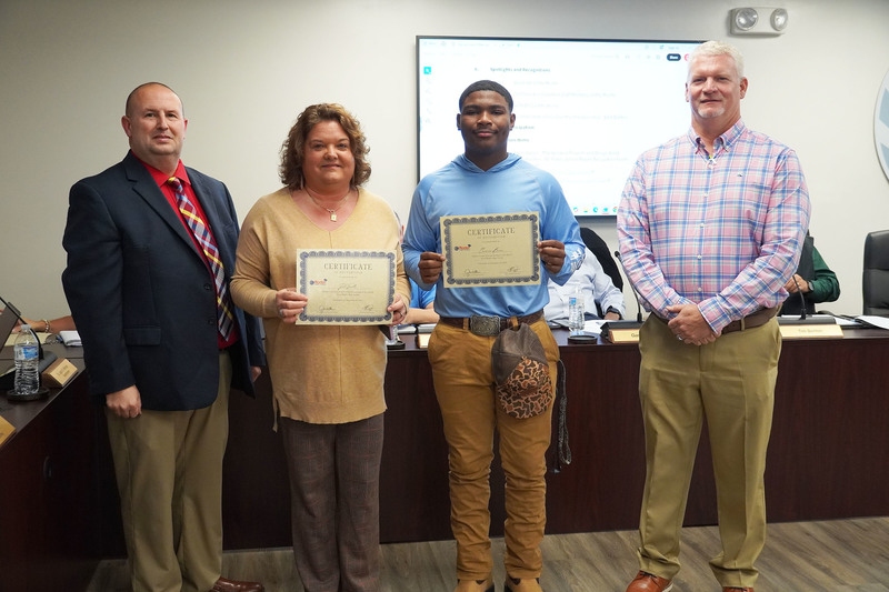 Staff member and student stand with certificates beside two administrators at a board meeting.