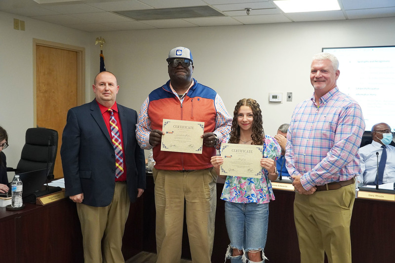 Two students hold certificates while standing between two adult administrators during a recognition at a Bladen County Schools meeting.