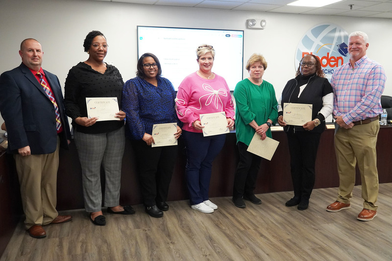 Six staff members stand in a row holding certificates at a Bladen County Schools board meeting, flanked by two administrators.