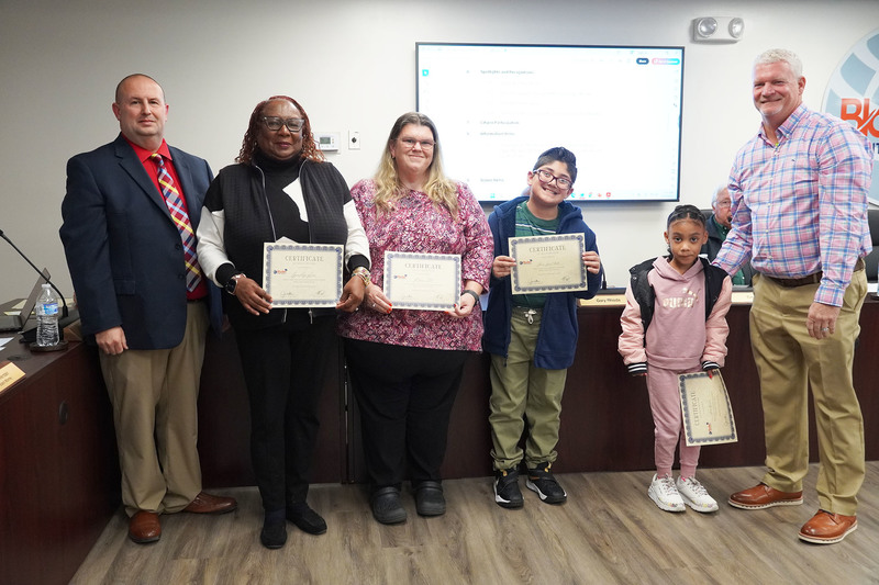 Two staff members and two students hold certificates alongside two administrators during a recognition at a board meeting.
