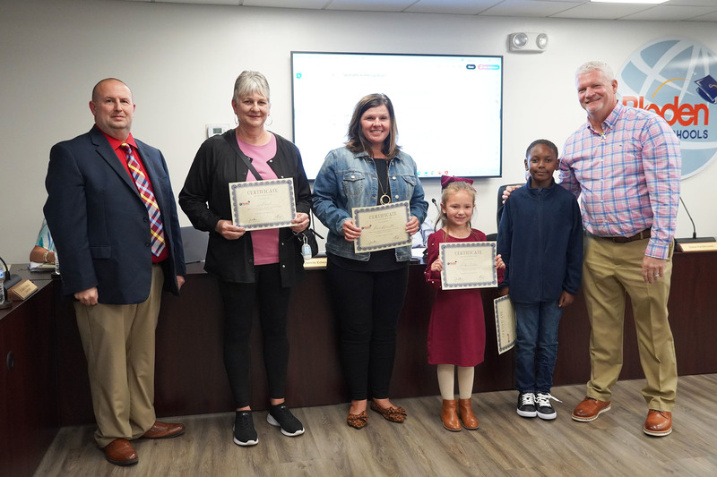 Two staff members and two students stand with certificates between two administrators at a Bladen County Schools board meeting.