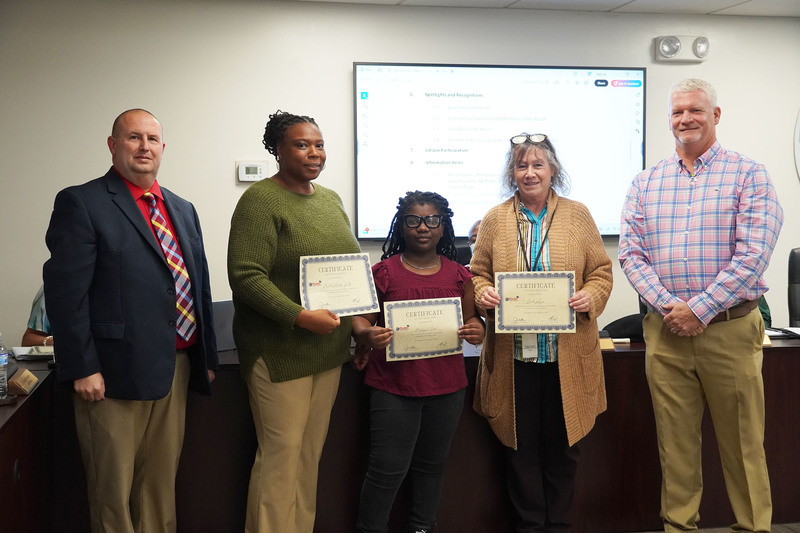 Two staff members and a student stand holding certificates with two administrators during a recognition at a Bladen County Schools meeting.