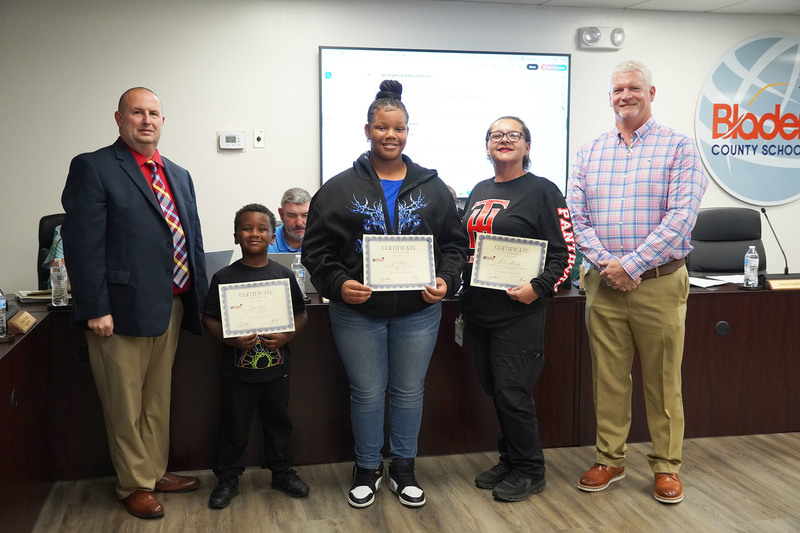 A young student, an older student, and a staff member hold certificates and stand with two administrators at a Bladen County Schools meeting.