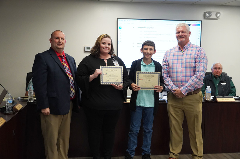 A staff member and a student hold certificates while standing between two administrators at a Bladen County Schools board meeting.