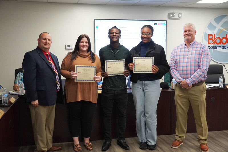 Two students and a staff member hold certificates and stand between two administrators at a Bladen County Schools meeting.