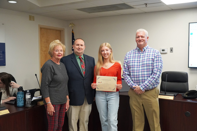 A student holds a certificate while standing with three adults in a boardroom. They face the camera for a recognition photo, with chairs, nameplates, and meeting materials visible in the background.