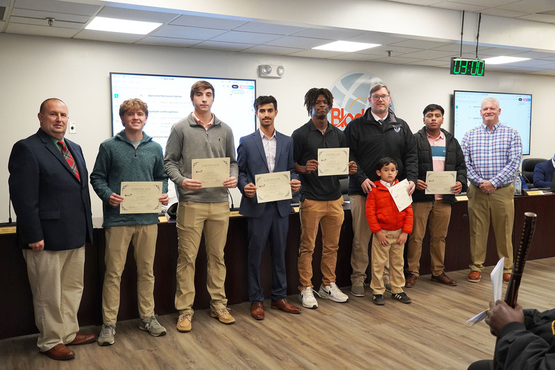 A group of students stands in a boardroom holding certificates, joined by two adults and a young child. They face the camera for a recognition photo, with meeting screens and nameplates visible in the background.