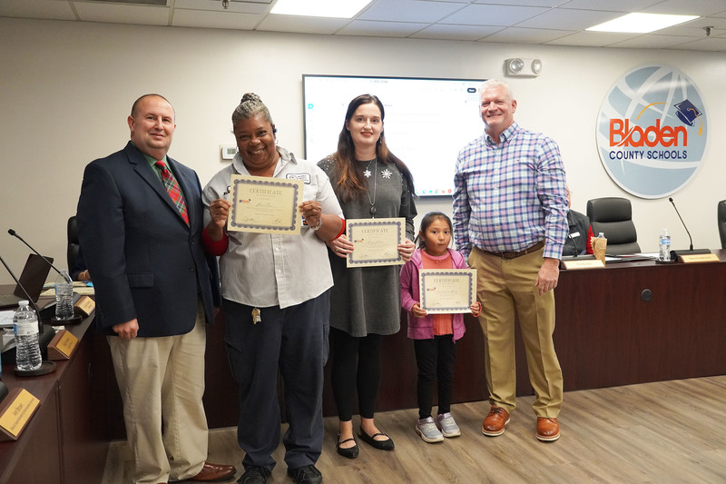 Two adults and a young student hold certificates while posing with two district leaders in a boardroom. A Bladen County Schools logo is visible on the wall behind them.
