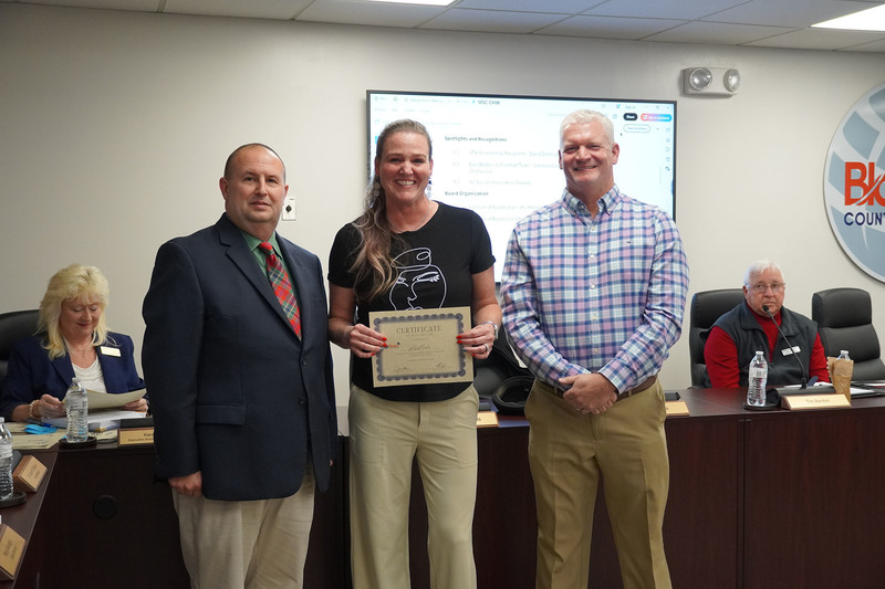 An adult award recipient stands in the center holding a certificate while posing with two district leaders in a boardroom setting.