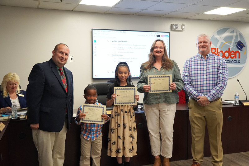 A young student, an older student, and an educator hold certificates while posing with two district leaders in a boardroom.