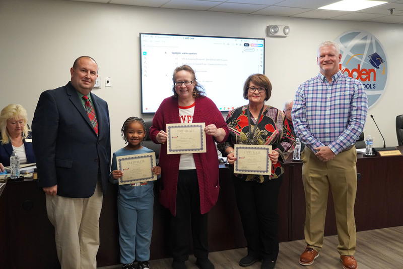 A young student, two adults, and a district leader pose with certificates in a boardroom with officials seated behind them.