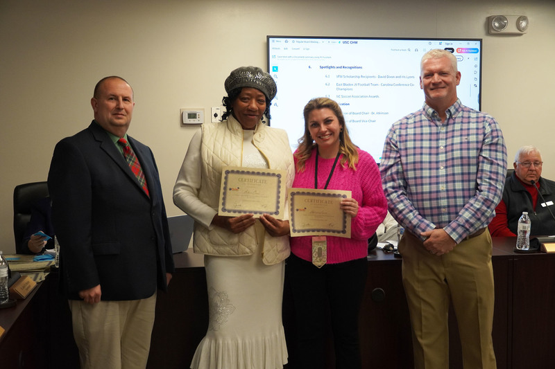 Two adult award recipients pose with certificates beside district leaders in a boardroom, smiling toward the camera.