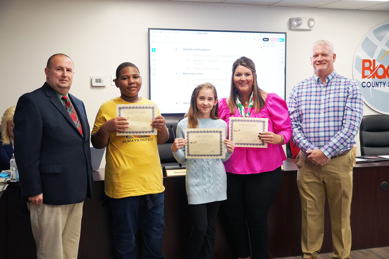 Two students and an educator hold certificates while posing with two district leaders in a boardroom. A meeting screen is visible behind them.