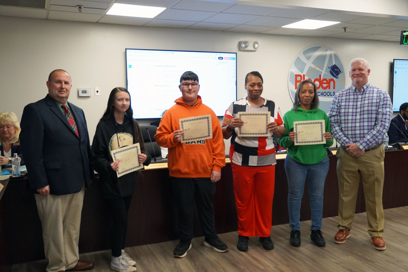 A group of four award recipients holds certificates while standing between two district leaders in a boardroom. Everyone faces the camera, with the Bladen County Schools logo visible behind them.