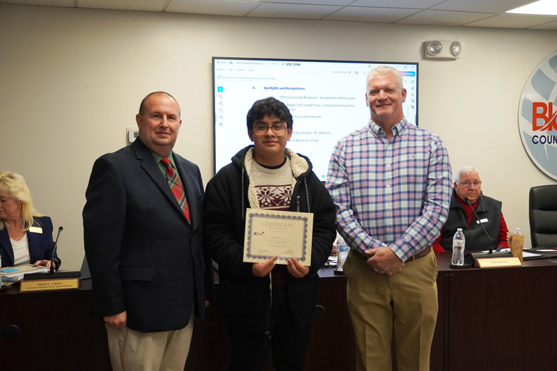 A student holds a certificate while standing with two adults in a boardroom. They smile at the camera with meeting screens and officials visible in the background.