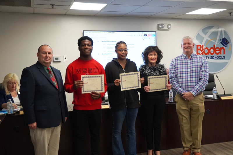 Three award recipients stand beside two district leaders holding certificates in a boardroom. Meeting materials and a Bladen County Schools logo appear in the background.