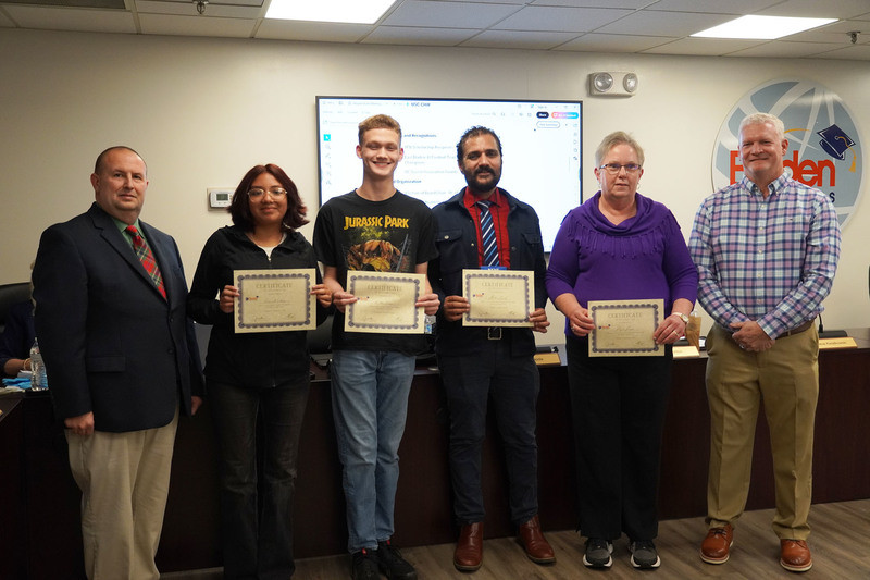 Four award recipients hold certificates and stand with district leaders in a boardroom, smiling for a recognition photo.