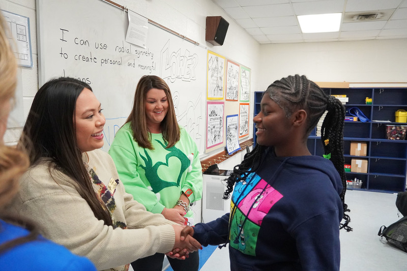 Teacher shakes hands with a student in a classroom while another staff member looks on, celebrating student engagement.