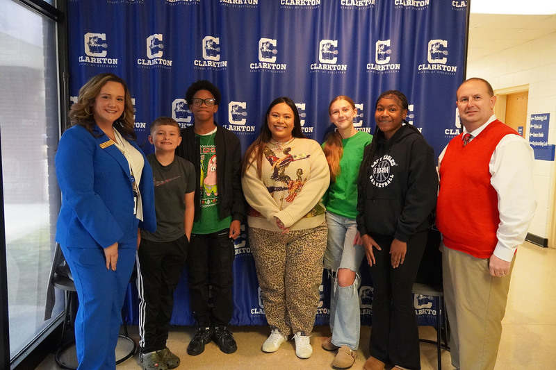 Seven people, including students and staff, stand smiling in front of a Clarkton School of Discovery step-and-repeat banner inside a school hallway.