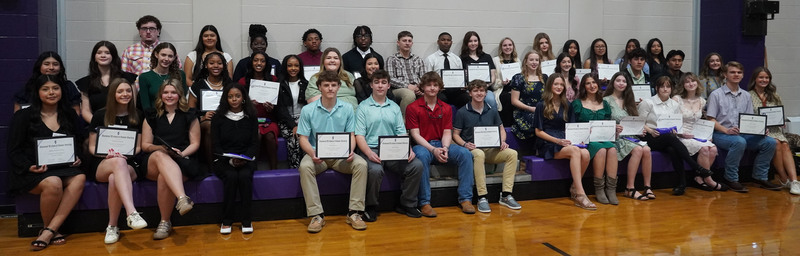 Large group of high school students seated on gym bleachers holding certificates during a recognition ceremony.