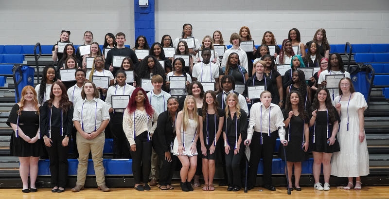 Large group of high school students standing on gym bleachers holding certificates and wearing honor cords at an awards event.