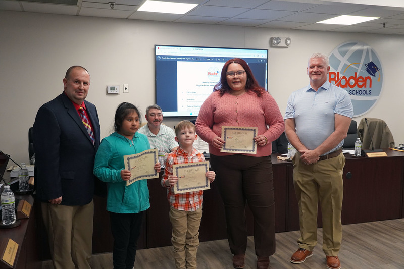 Two elementary students and an adult recipient pose with district leaders while holding certificates of recognition in the Bladen County Schools boardroom.