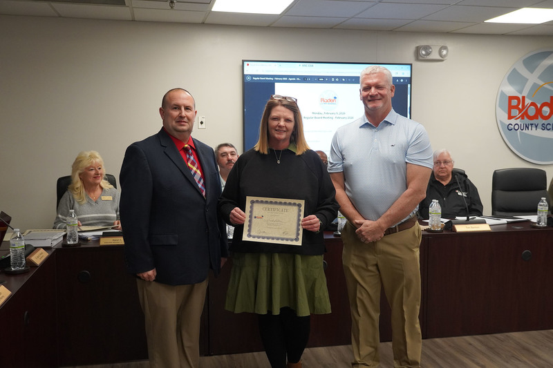 An educator stands between district leaders, holding a certificate of recognition during a Bladen County Schools Board of Education meeting, with board members seated in the background.