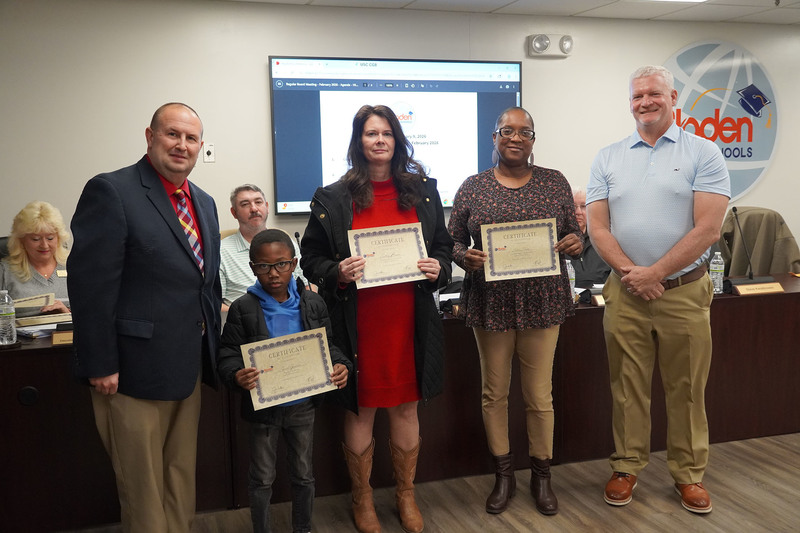 Three certificate recipients, including a student and two adults, pose with district leaders while holding certificates of recognition at a Bladen County Schools Board of Education meeting.