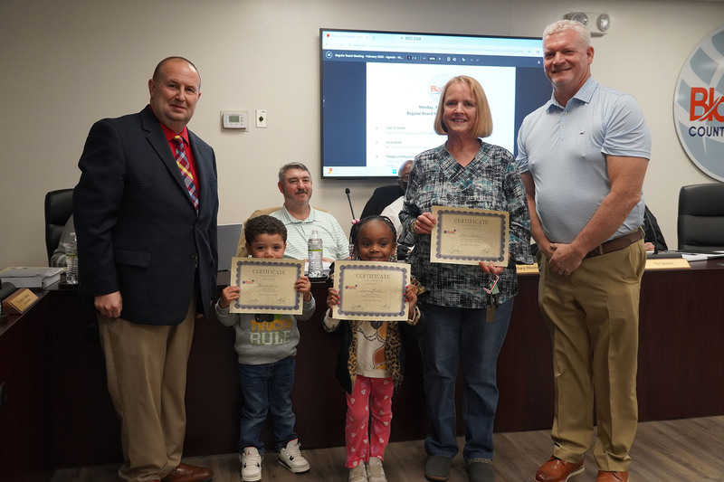 Two elementary-aged students and an adult stand with district leaders, holding certificates of recognition in the Bladen County Schools boardroom.