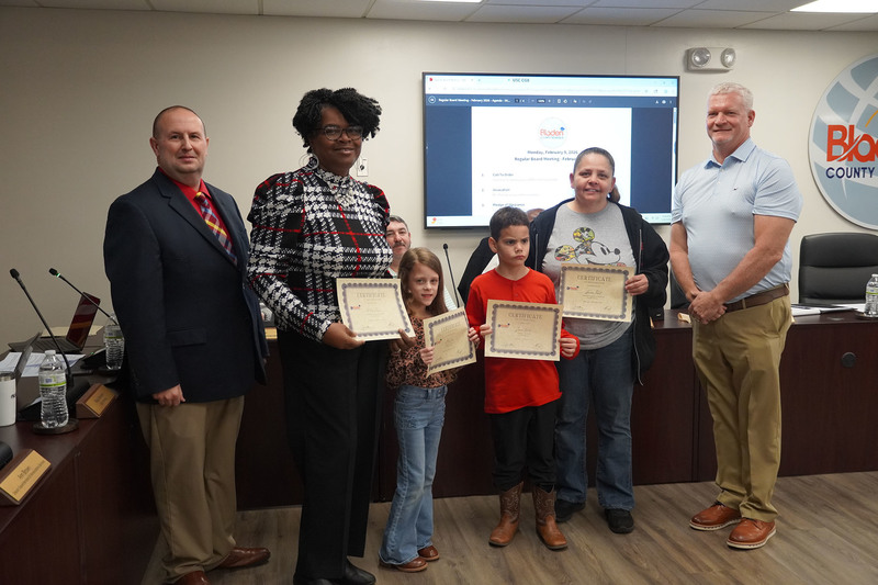 An educator and two students pose with district leaders while holding certificates of recognition at a Bladen County Schools Board of Education meeting.