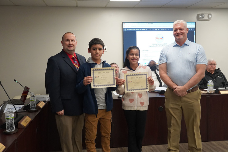 Two students stand between district leaders, each holding a certificate of recognition during a Bladen County Schools Board of Education meeting.